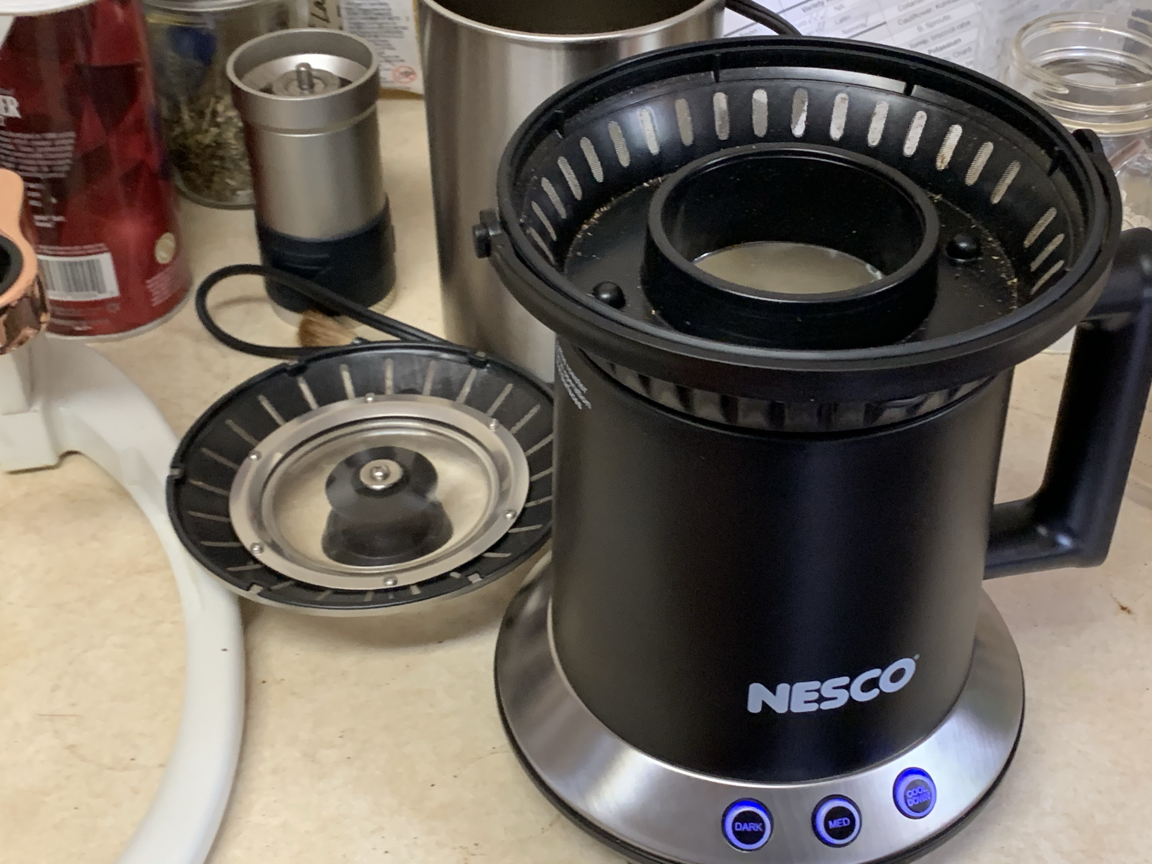 Image of Nesco coffee roaster with lid removed in forground, multiple items in background including Flair Espresso maker, kosher salt shaker, and manual coffee grinder.