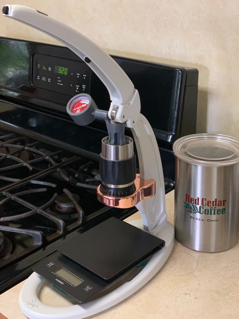 Picture of Flair Espresso Maker, with Hario scale, Red Cedar Airscape Coffee canister, and a gas stove in the background.